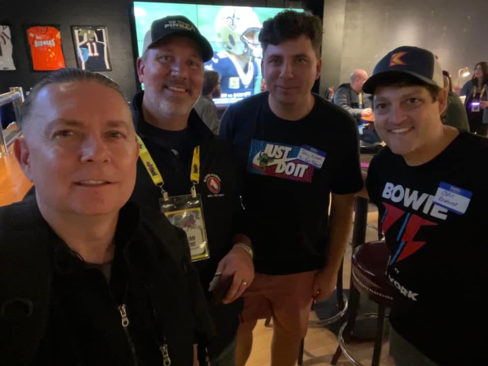 Four men at event wearing caps and shirts, smiling together