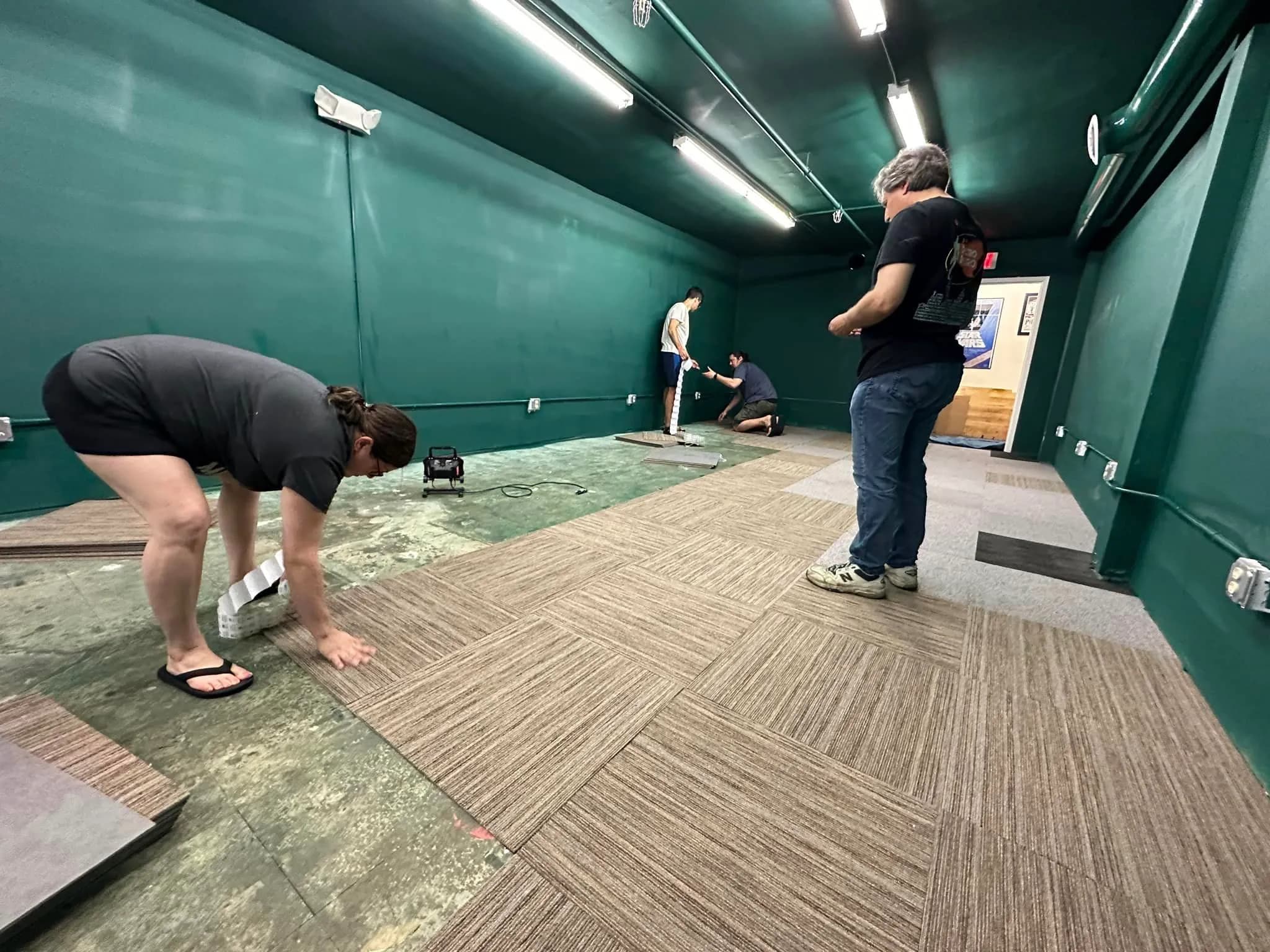 Workers installing carpet tiles in a green-walled room with fluorescent lighting