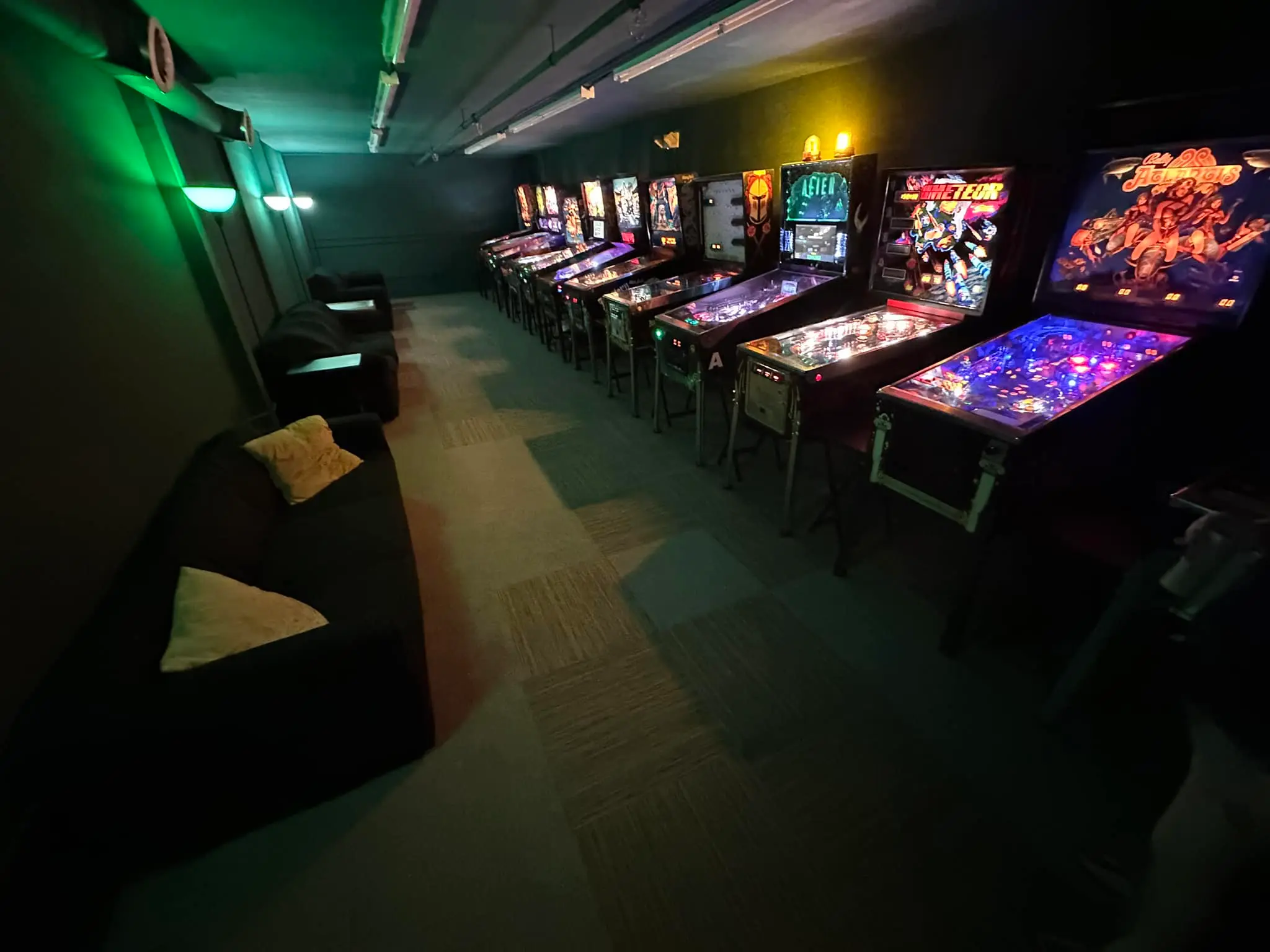 Dimly lit arcade room with colorful pinball machines and green lighting