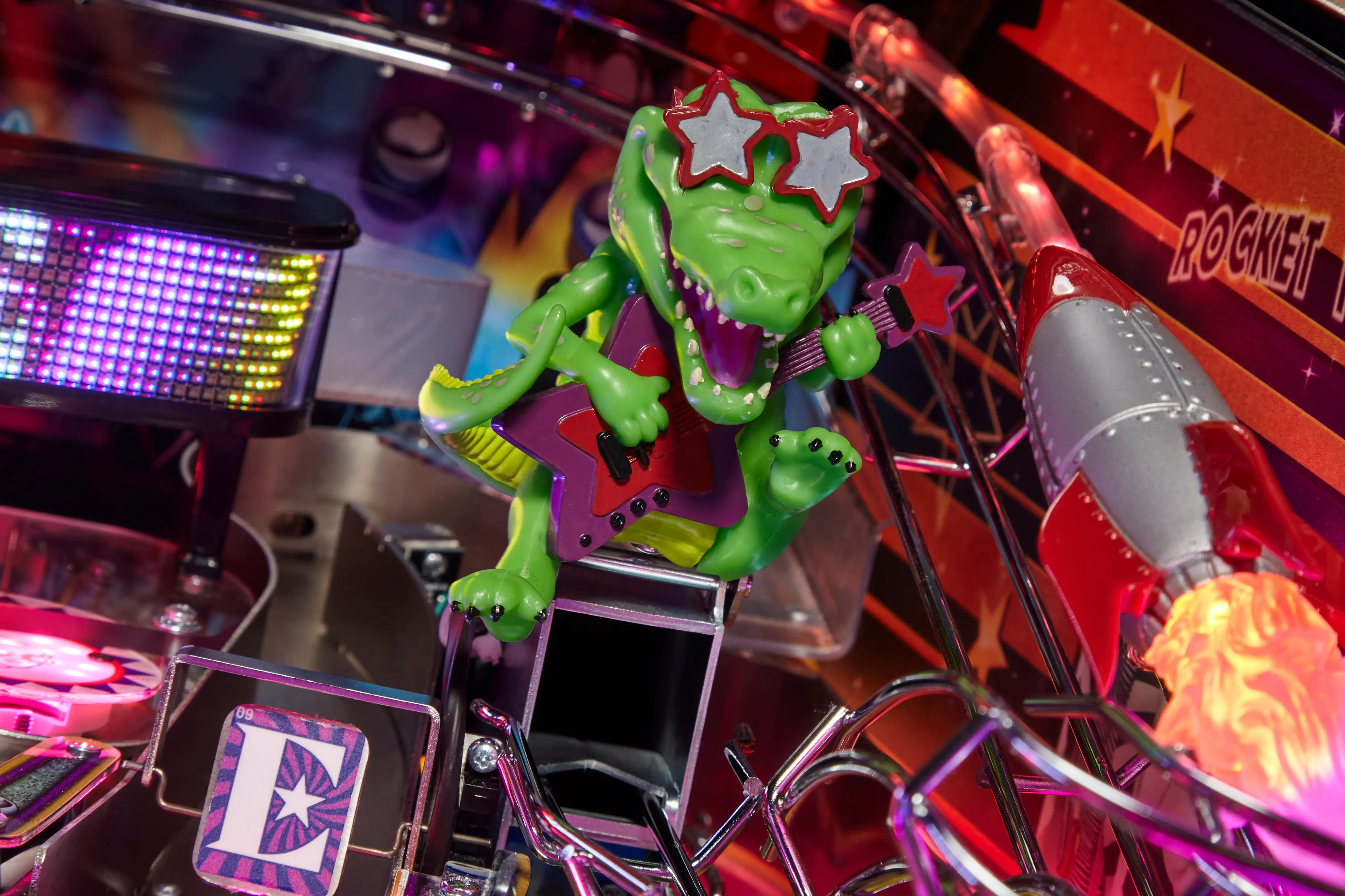 Green alien rocker with star sunglasses poses on colorful pinball machine