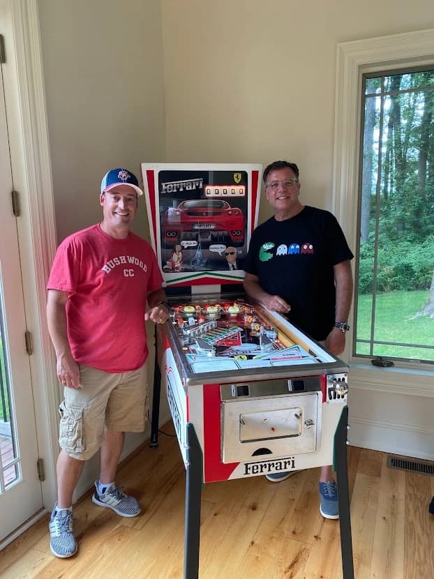 Two men posing with a Ferrari-themed pinball machine at home