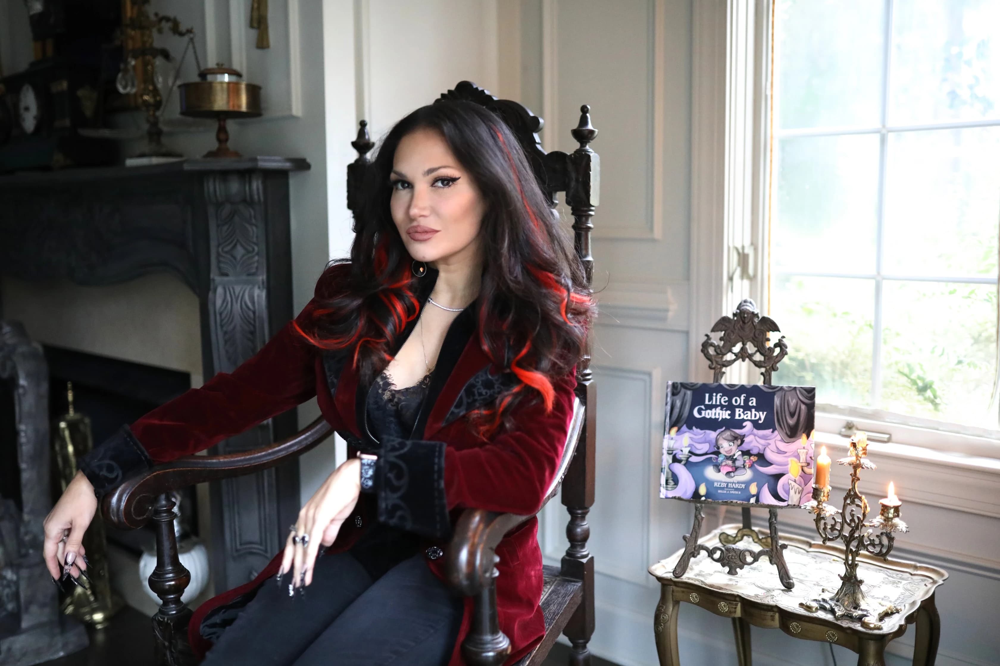 Gothic-styled woman in red jacket sitting by fireplace with book and candelabra