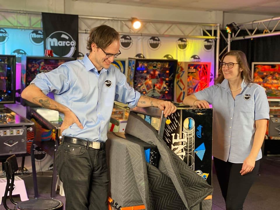 Two employees at Marc arcade showcase colorful pinball machines and stage backdrop